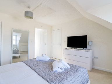 A bedroom with a bed, rolled towels, a dresser with a TV, a floor mirror, and white doors at The Ferns in Weymouth