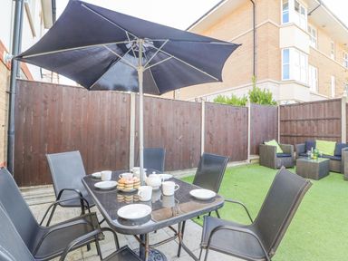 An outdoor patio area with a table set with cups plates and cupcakes under a large umbrella and seating with cushions on artificial grass at The Ferns in Weymouth
