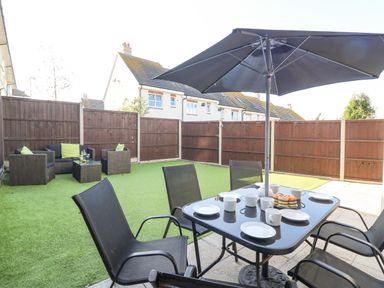 An outdoor patio area with a dining table and chairs under an umbrella and seating area with sofas and a coffee table on artificial grass at The Ferns in Weymouth