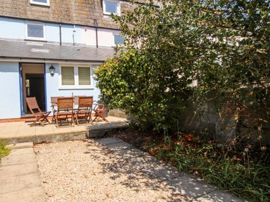 An outdoor patio area with wooden chairs and a table next to a blue and white house with a tree and garden bed in the foreground at Firkin Cottage Brewers Quay Harbour