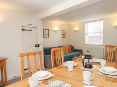 A dining area with wooden table and chairs set with white dishes and mugs next to a living area with a sofa and cushions at Firkin Cottage in Brewers Quay Harbour