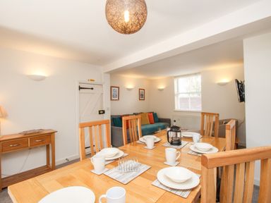 A dining table set with white dishes and mugs in a dining room with a wooden side table and a couch with colorful cushions at Firkin Cottage in Brewers Quay Harbour