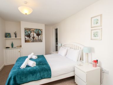 A bedroom with a white bed and teal blanket with rolled towels on it two bedside tables with lamps and wall art at Firkin Cottage Brewers Quay Harbour