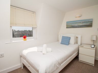 A bedroom with a single bed with white linens and a blue striped pillow beside a window and a nightstand with a lamp and clock at Firkin Cottage Brewers Quay Harbour