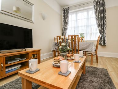 A living room with a wooden coffee table set with cups and muffins a wooden dining table with chairs against a window with curtains at Footprint in Brewers Quay Harbour