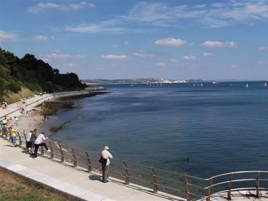 A coastal walkway with people looking over a railing at the sea with a beach on the left and distant hills at Georges House in Brewers Quay Harbour