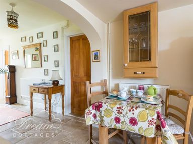 A dining area with a table covered by a floral tablecloth set with cups and scones wooden chairs a wooden wall cabinet and a small wooden side table with a lamp at Grazeland Cottage in Bridport