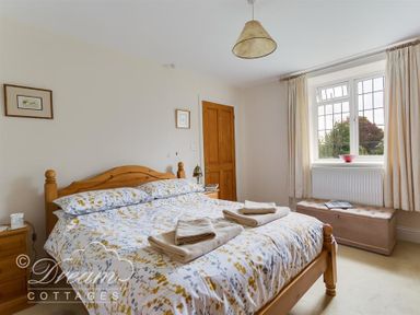 A bedroom with a wooden double bed a bedside table a window with curtains and a bench at Grazeland Cottage in Bridport