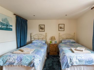 A bedroom with two single beds separated by a wooden nightstand with a lamp and two cups at Grazeland Cottage in Bridport