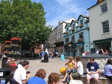 An outdoor public square with people sitting and walking near buildings with flower decorations at Grazeland Cottage in Bridport