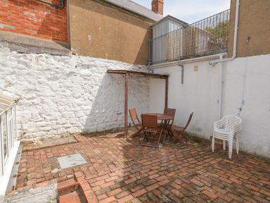 An outdoor brick patio area with a wooden table and four chairs stacked plastic chairs and a white painted brick wall at Guinea Cottage in Brewers Quay Harbour