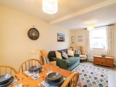 A living room with a wooden dining table and chairs a green sofa beige armchairs a clock on the wall and a window with curtains at Guinea Cottage in Brewers Quay Harbour