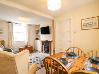 A living room and dining area with a wooden dining table set with bowls and glasses a fireplace mounted tv armchairs a window with curtains and a rug on the floor at Guinea Cottage Brewers Quay Harbour