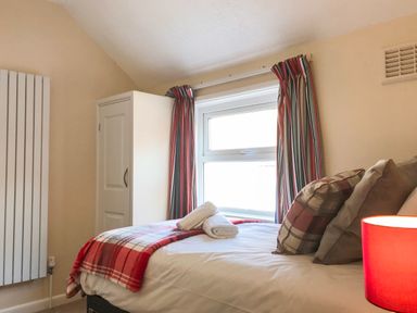 A bedroom with a bed covered with white linens and a red plaid blanket two rolled white towels on the bed striped curtains on the window a white wardrobe and a red lampshade near the bed at Guinea Cottage Brewers Quay Harbour
