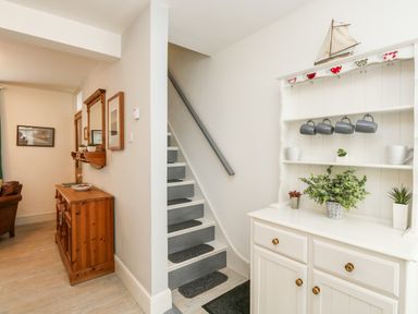 A hallway with a white cabinet holding mugs and plants next to a staircase and a wooden sideboard with a mirror at Harbour Catch Cottage in Weymouth