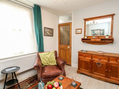 A living room with a brown leather armchair and green cushion a wooden cabinet with a mirror above and a wooden table with fruit and bottles at Harbour Catch Cottage in Weymouth