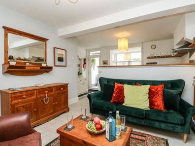A living room with a green sofa with red and green cushions a wooden coffee table with bottles and a fruit bowl and a wooden sideboard with a mirror at Harbour Catch Cottage in Weymouth