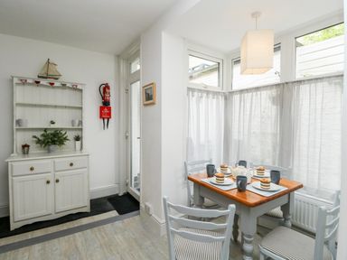 A dining area with a wooden table set for four by windows with sheer curtains and a white cabinet with plants at Harbour Catch Cottage in Weymouth