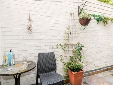 An outdoor patio with a round mosaic table holding bottles and glasses a black chair a potted plant climbing a trellis and a hanging basket on a white brick wall at Harbour Catch Cottage in Weymouth