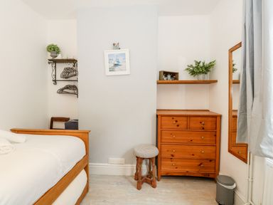 A bedroom with a wooden bed, chest of drawers, stool, wall-mounted shelves with hangers and plants, and a large wall mirror at Harbour Catch Cottage in Weymouth