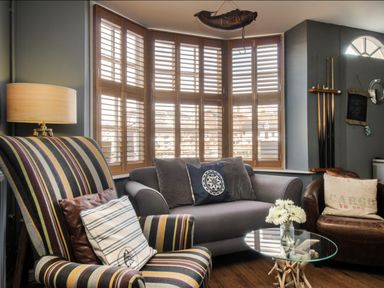 A sitting area with a striped armchair, gray sofa, round glass coffee table, bedside lamp, and wooden window shutters at Harbourside House in Brewers Quay Harbour