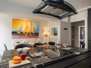 A kitchen island with a cooktop and bell peppers on a cutting board with a dining table and colorful sailboat painting in the background at Harbourside House in Brewers Quay Harbour