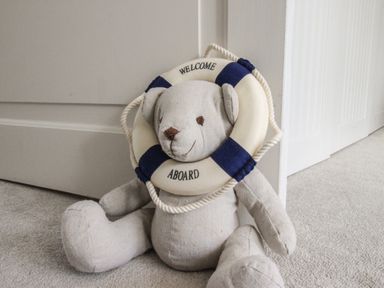 A teddy bear with a lifebuoy that says welcome aboard sitting on carpeted floor by a white door at Harbourside House in Brewers Quay Harbour