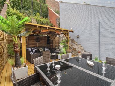 A patio area with a glass table and wicker chairs under a wooden pergola with seating and plants at Harbourside House in Brewers Quay Harbour