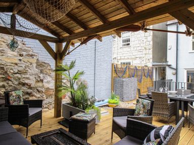 An outdoor patio area with wicker chairs and tables under a wooden pergola with plants and a stone wall at Harbourside House in Brewers Quay Harbour