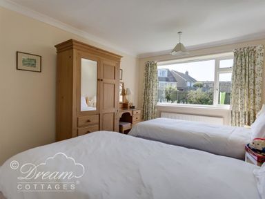 A bedroom with two single beds a wooden wardrobe a dressing table and a large window with floral curtains at Hillview Bungalow in Burton Bradstock