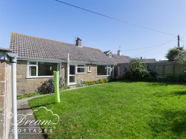 A backyard with green grass and bushes behind a single story house at Hillview Bungalow in Burton Bradstock