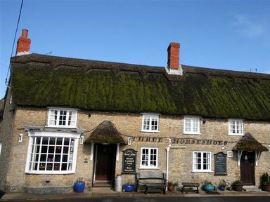 A stone building with a thatched roof and multiple windows with signs and pots outside at Three Horseshoes Burton Bradstock