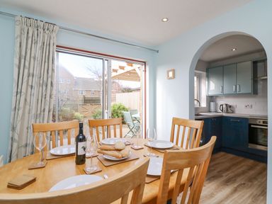 A dining area with a wooden table set with plates glasses and bread next to a window with curtains and an open view into a kitchen at Home Waters in Wyke Regis