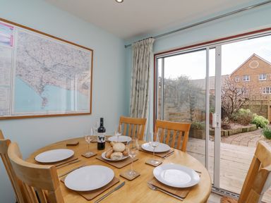 A dining area with a wooden table and chairs set with plates wine glasses and bread beside sliding glass doors overlooking a garden at Home Waters in Wyke Regis