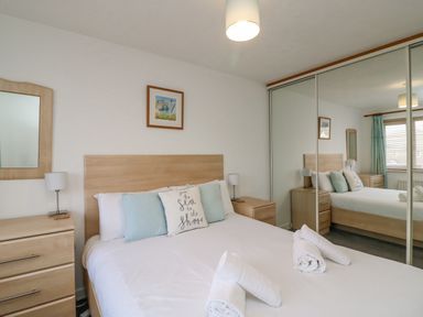 A bedroom with a double bed with pillows and rolled towels bedside tables with lamps a wooden dresser a wall mirror and mirrored sliding wardrobe doors at Home Waters in Wyke Regis