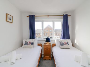 A bedroom with two single beds with white bedding and pillows with seaside house prints a wooden nightstand with a lamp is between the beds at Home Waters Wyke Regis