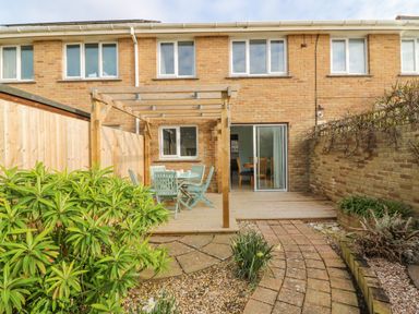 A backyard with a wooden pergola and outdoor dining set a brick-paved path and plants at Home Waters in Wyke Regis