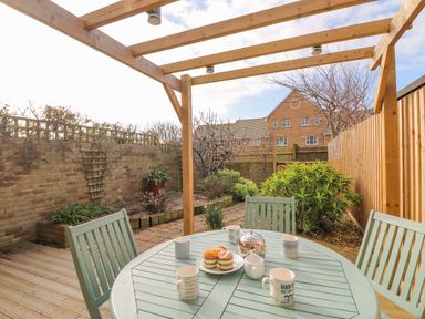 A patio area with a green table and chairs under a wooden pergola and a garden with plants and trees at Home Waters in Wyke Regis