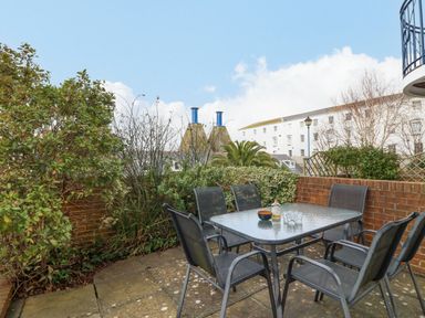 An outdoor patio with a glass table and six chairs surrounded by plants and brick walls at Hops House in Brewers Quay Harbour