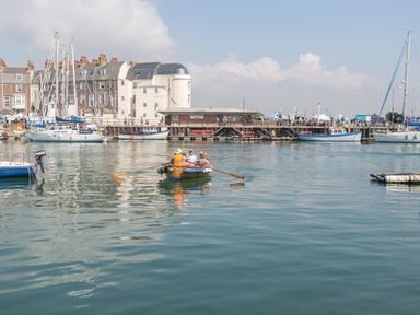 A small group of people rowing a wooden boat in a harbor with sailboats docked and buildings in the background at Brewers Quay Harbour