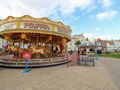 A carousel with people riding horses near a kiosk and benches in an outdoor urban area