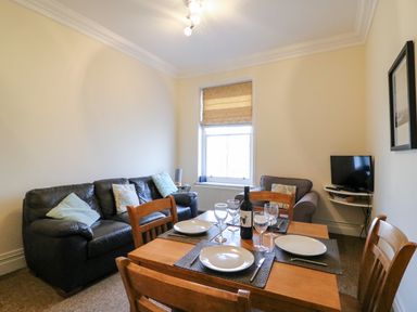 A dining table with plates and wine glasses beside a black sofa and an armchair with a television on a wall shelf at Harbour Watch Apartment 6 Brewers Quay Harbour