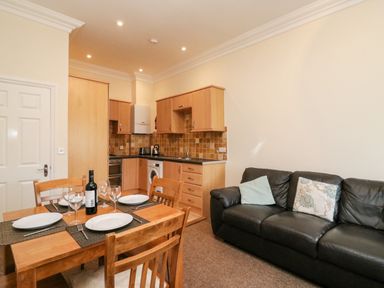A kitchen and dining area with wooden cabinets and a table set with plates and glasses next to a black sofa at Harbour Watch Apartment 6 in Brewers Quay Harbour
