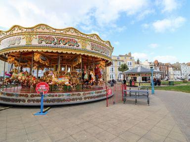 A carousel with people riding horses near a food kiosk and benches in an open public space