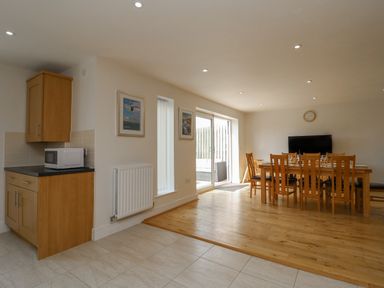 A dining area with a wooden table and chairs a microwave on a kitchen counter and a television mounted on the wall at Jurassic Coast House in Portland
