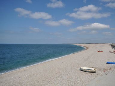 A pebble beach with small boats on the shore and a blue sea under a partly cloudy sky at Langham Cottage in Portland