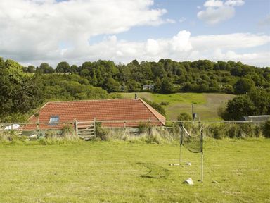 A grassy field with a small badminton net and a building with a red tiled roof with trees and hills in the background at Leys At Valley View Farm in Uplyme