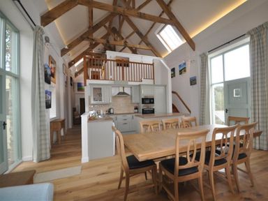A dining area with wooden chairs and table next to a kitchen with cabinets and appliances under a vaulted ceiling with exposed beams at Leys At Valley View Farm in Uplyme