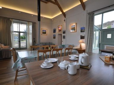 A dining and living area with wooden chairs and tables teapot and cups on the table paintings on the wall and large windows at Leys At Valley View Farm in Uplyme