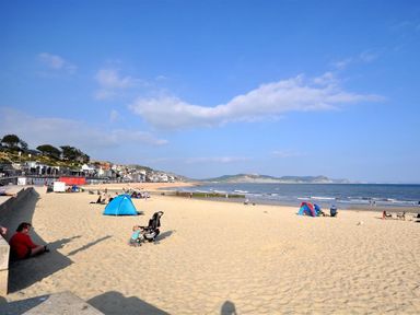 A sandy beach with people sitting and small tents near the shoreline with houses and hills in the background at Leys At Valley View Farm in Uplyme